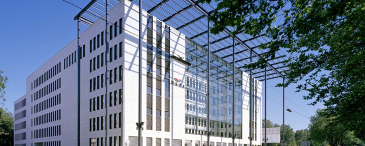 Modernes Bürogebäude mit Glasfassade und umliegender grüner Landschaft, unter blauem Himmel.