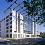 Modernes Bürogebäude mit Glasfassade und umliegender grüner Landschaft, unter blauem Himmel.