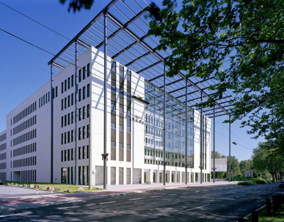 Modernes Bürogebäude mit Glasfassade und umliegender grüner Landschaft, unter blauem Himmel.