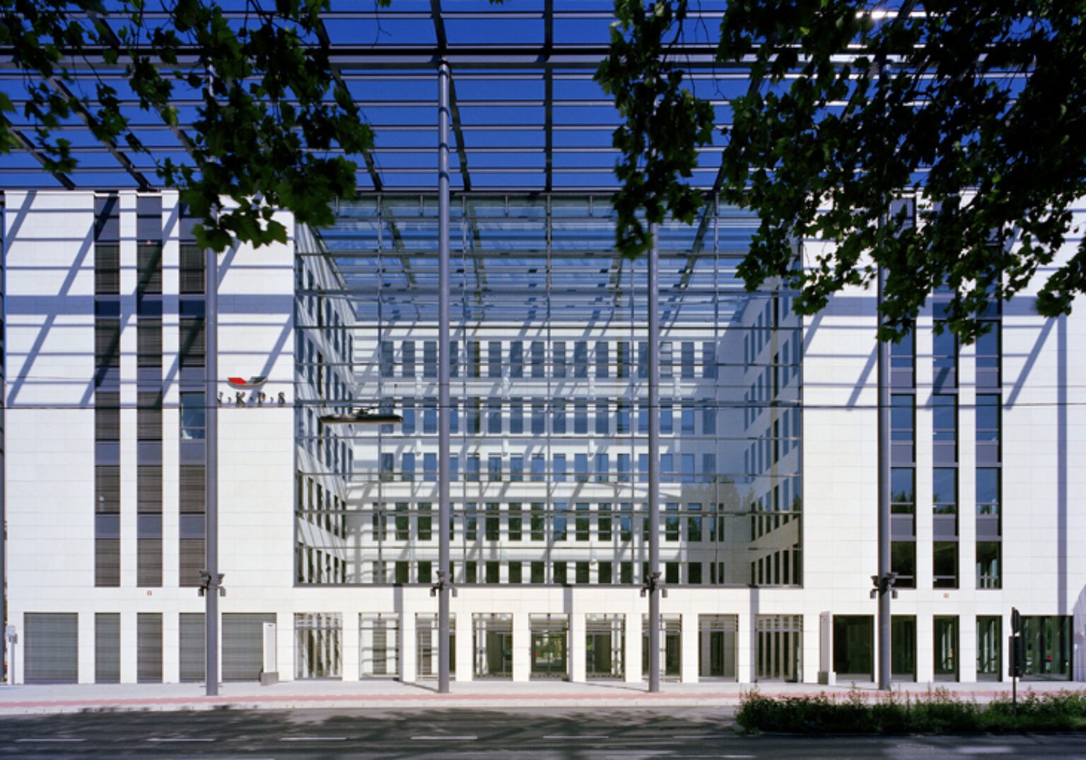 Moderne Glasfassade eines Bürogebäudes bei klarem Himmel, umgeben von Bäumen und Spiegelungselementen.