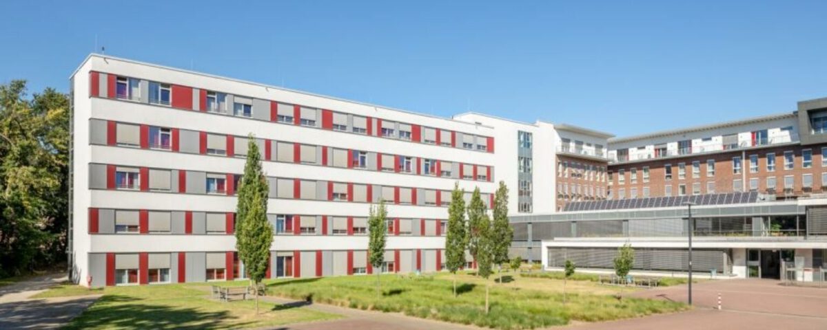 Modernes Krankenhausgebäude mit rot-weißer Fassade und grüner Landschaft bei klarem Himmel.