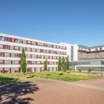 Modernes Krankenhausgebäude mit rot-weißer Fassade und grüner Landschaft bei klarem Himmel.