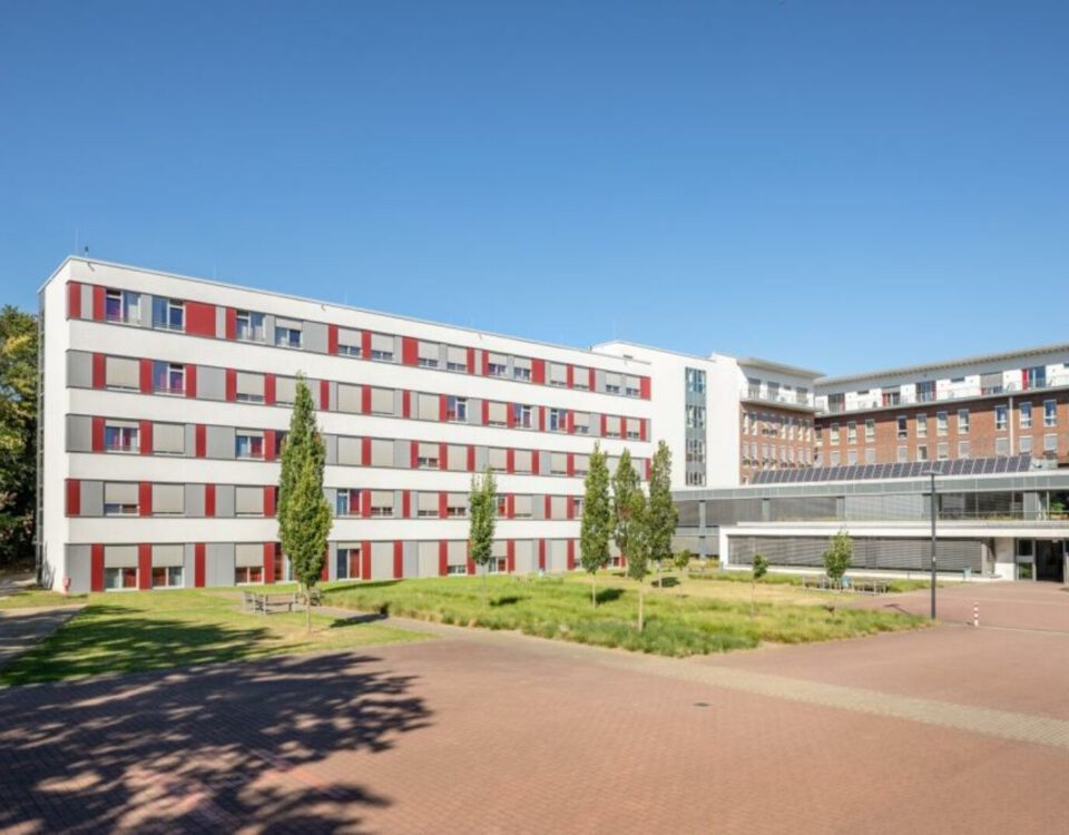 Modernes Krankenhausgebäude mit rot-weißer Fassade und grüner Landschaft bei klarem Himmel.