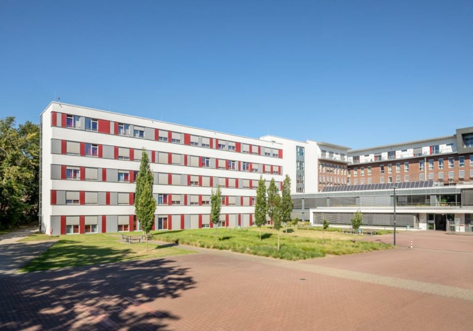 Modernes Krankenhausgebäude mit rot-weißer Fassade und grüner Landschaft bei klarem Himmel.