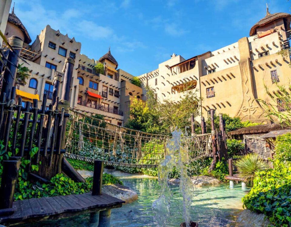 Afrika-inspiriertes Hotel mit Hängebrücke und Wasserfall im sonnigen grünen Garten, perfekter Urlaubsort für Familien.