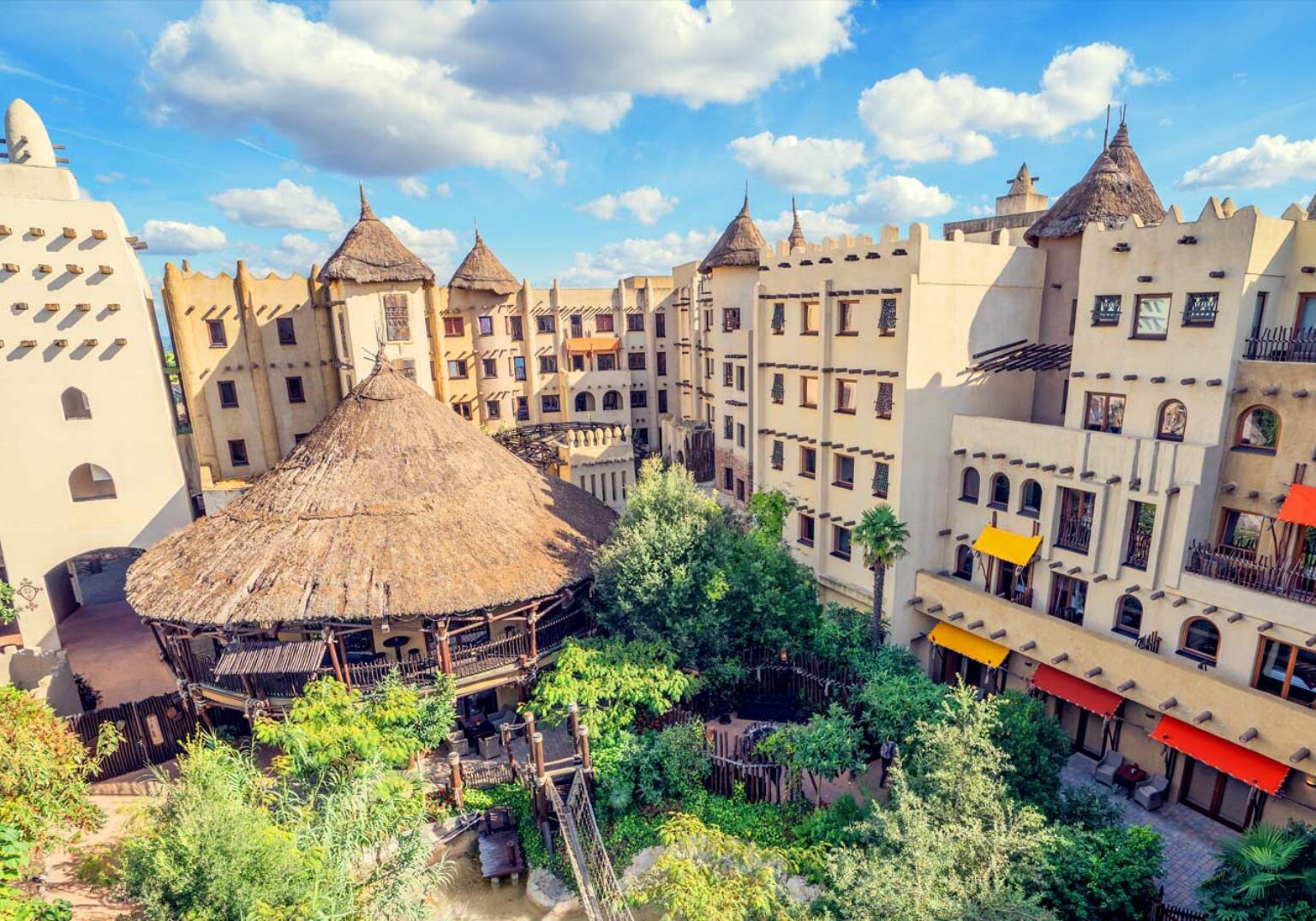 Afrikanisch inspiriertes Hotel mit Strohdach und Palmen in sonniger Umgebung, blauem Himmel und malerischer Architektur.