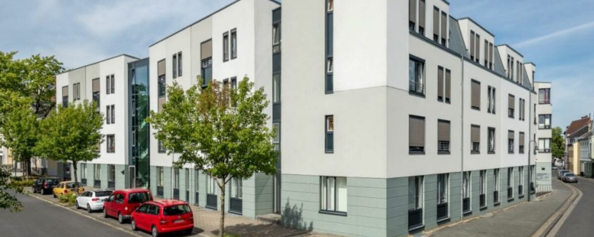 Moderne Wohnanlage an einer ruhigen Straßenecke mit geparkten Autos und begrünten Bäumen, bei sonnigem Wetter.