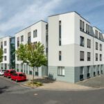 Moderne Wohnanlage an einer ruhigen Straßenecke mit geparkten Autos und begrünten Bäumen, bei sonnigem Wetter.