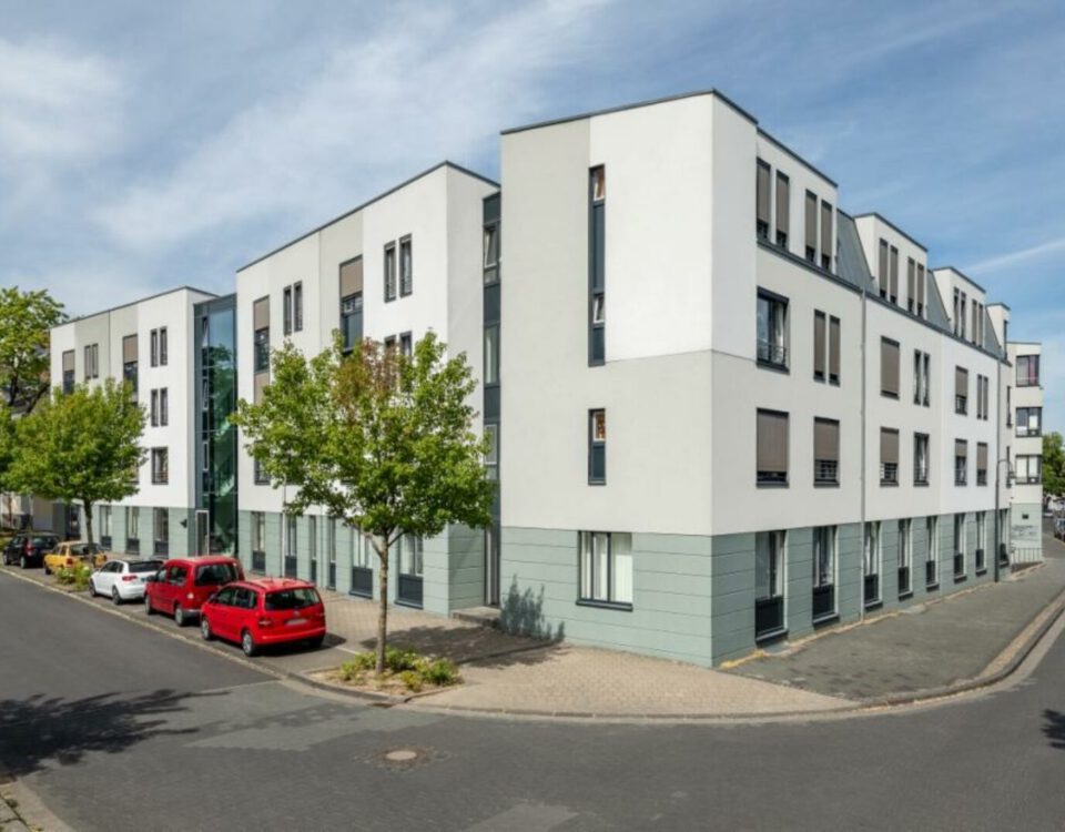 Moderne Wohnanlage an einer ruhigen Straßenecke mit geparkten Autos und begrünten Bäumen, bei sonnigem Wetter.