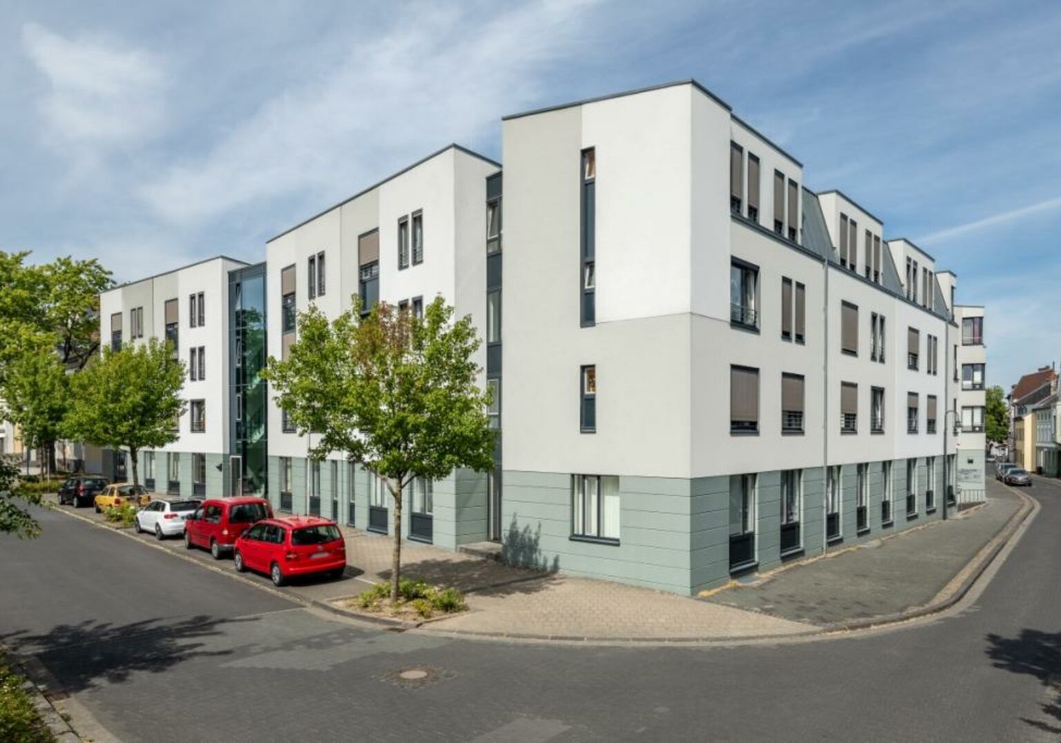 Moderne Wohnanlage an einer ruhigen Straßenecke mit geparkten Autos und begrünten Bäumen, bei sonnigem Wetter.