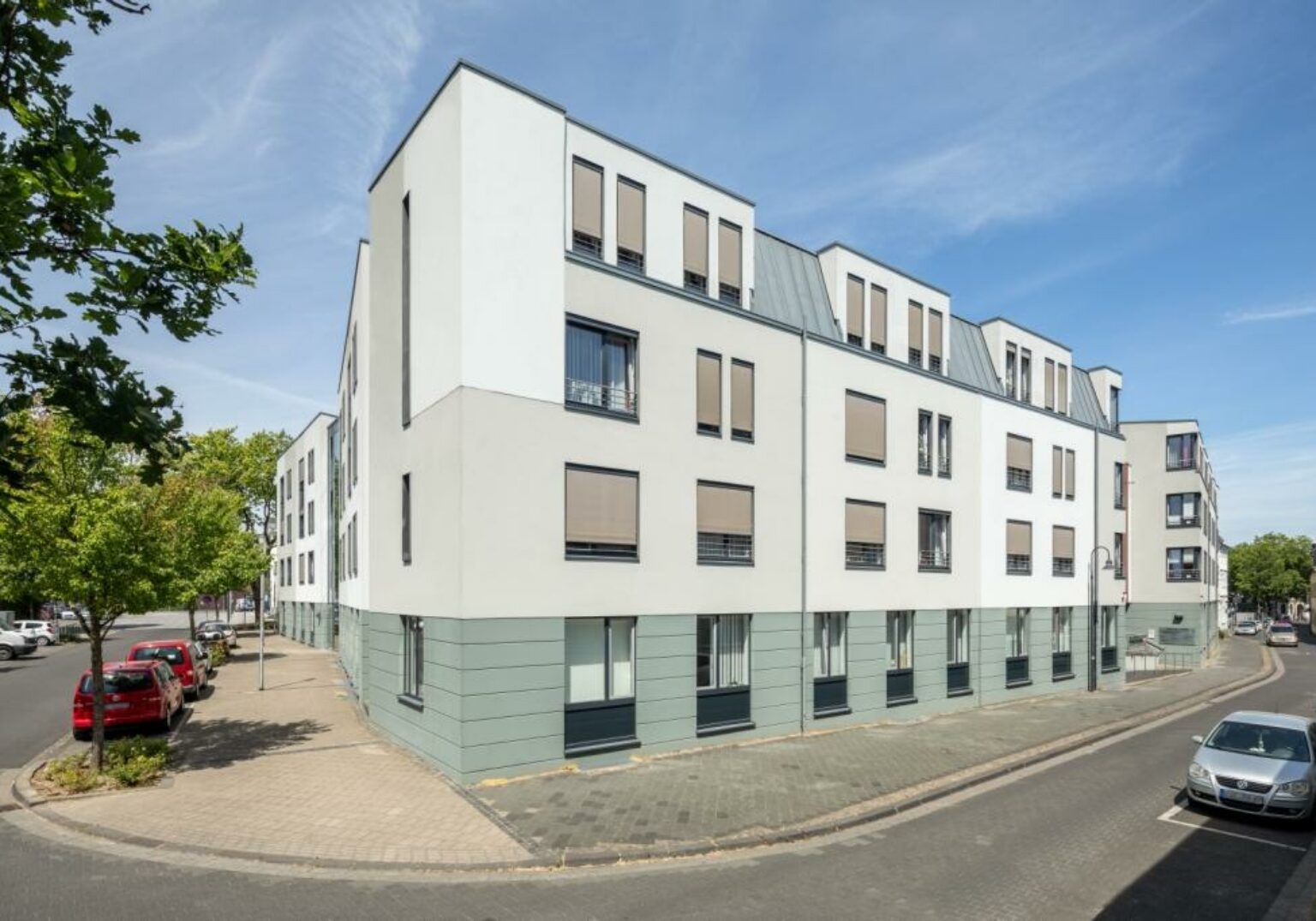 400212_Haus_Caritas_2_klein-1536x1075.2-c-default Moderne Mehrfamilienhausfassade an sonniger Straßenecke mit geparkten Autos und blauem Himmel.