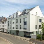 Moderne Wohngebäude an einer ruhigen Stadtstraße mit blauen Fensterelementen und grünen Akzenten.