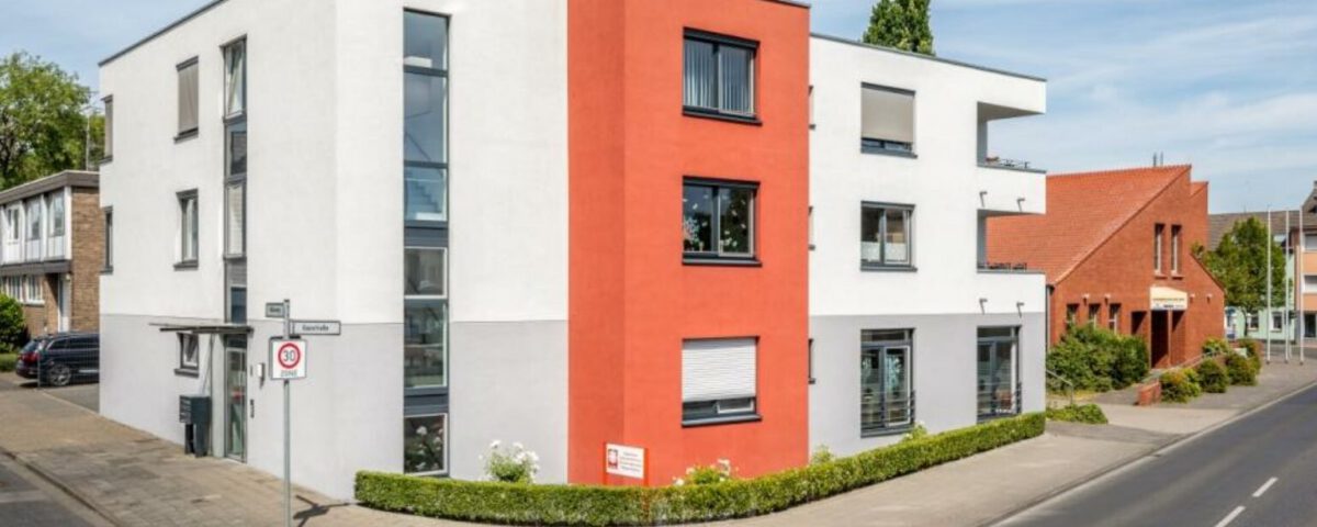 Modernes Wohngebäude mit roter Fassade an einer Straßenecke im Stadtgebiet, klare Architektur und blauer Himmel.