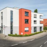 Modernes Wohngebäude mit roter Fassade an einer Straßenecke im Stadtgebiet, klare Architektur und blauer Himmel.