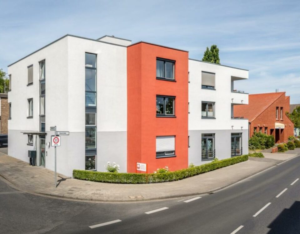 Modernes Wohngebäude mit roter Fassade an einer Straßenecke im Stadtgebiet, klare Architektur und blauer Himmel.