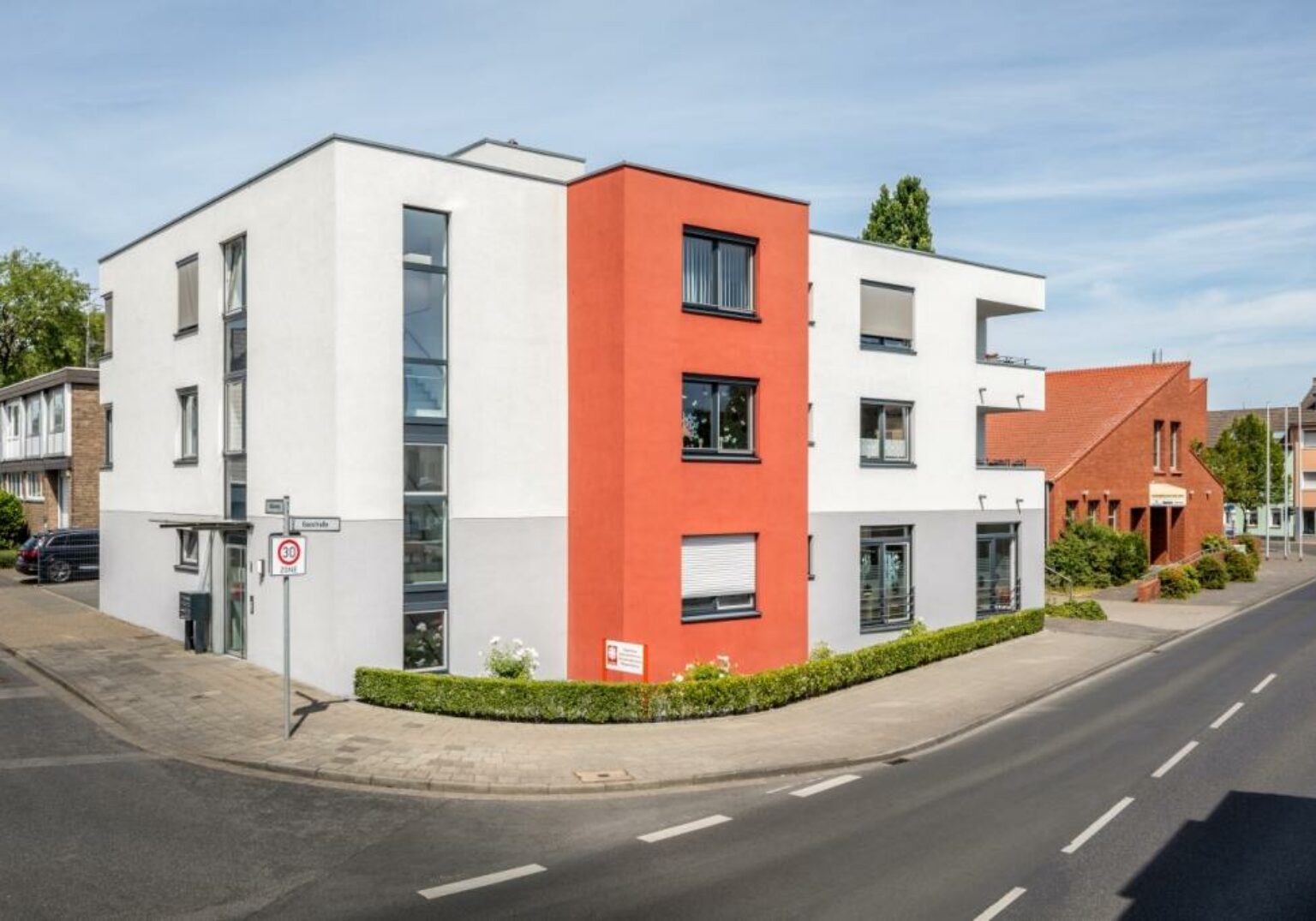 Modernes Wohngebäude mit roter Fassade an einer Straßenecke im Stadtgebiet, klare Architektur und blauer Himmel.