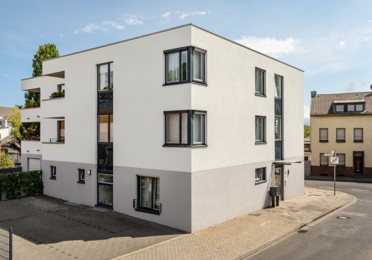 Modernes Wohngebäude an Straßenecke mit großen Fenstern und hellem Putz, sonniger Tag.