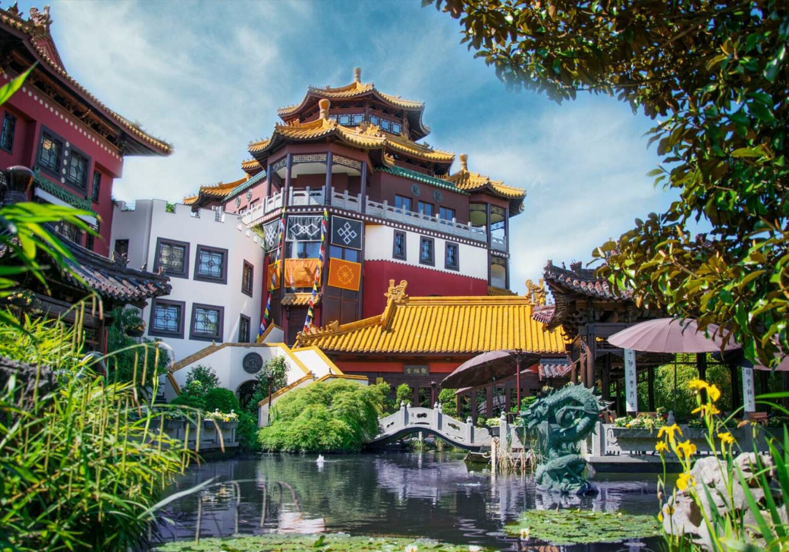 Chinesischer Pavillon mit Teich und Gärten im Vordergrund, strahlende Farben und traditionelle Architektur unter blauem Himmel.