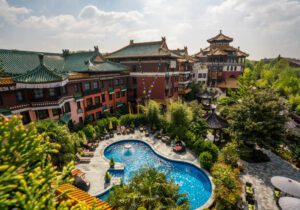 Asiatisch inspiriertes Hotel mit Pool und üppigen Gärten, umgeben von traditionellen Gebäuden an einem sonnigen Tag.