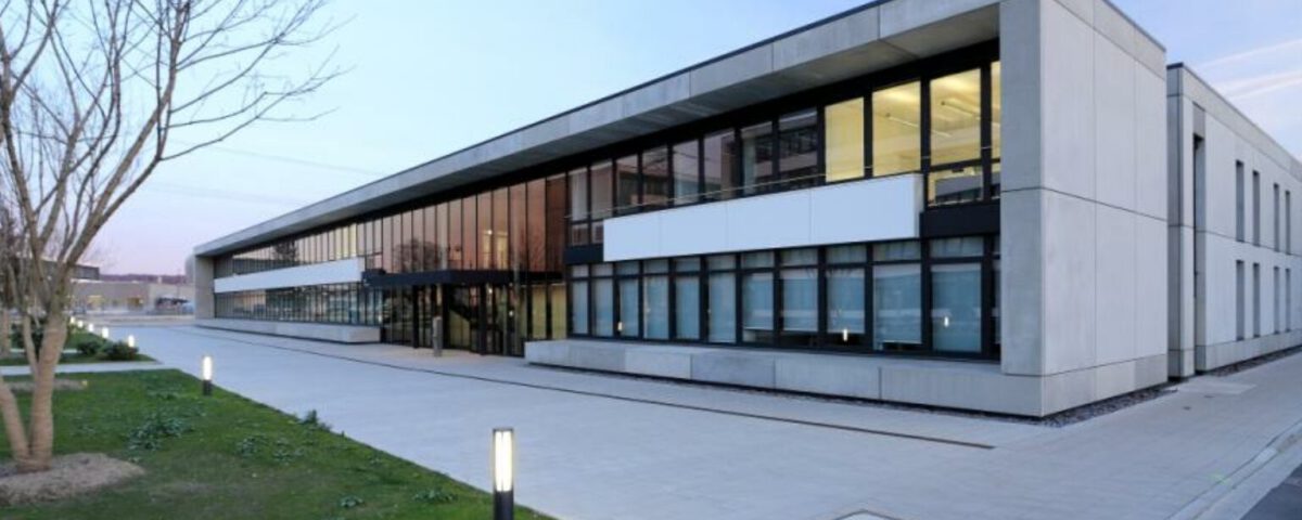 Moderne Bürogebäude-Fassade bei Sonnenuntergang mit Glasfenstern und gepflastertem Eingang, umgeben von Grünflächen.