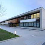 Moderne Bürogebäude-Fassade bei Sonnenuntergang mit Glasfenstern und gepflastertem Eingang, umgeben von Grünflächen.