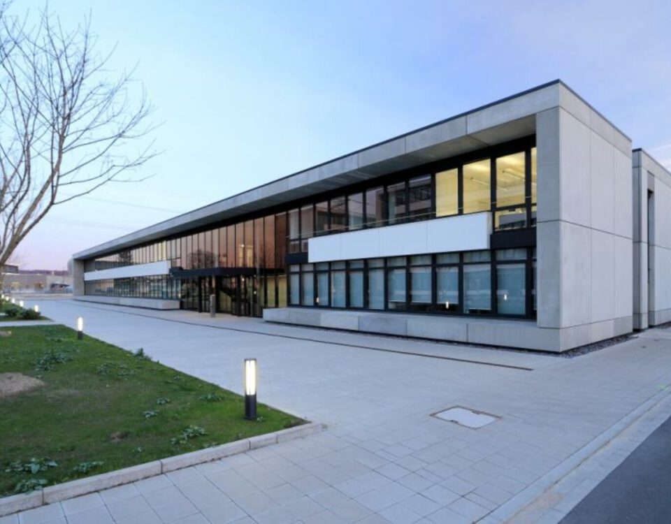 Moderne Bürogebäude-Fassade bei Sonnenuntergang mit Glasfenstern und gepflastertem Eingang, umgeben von Grünflächen.
