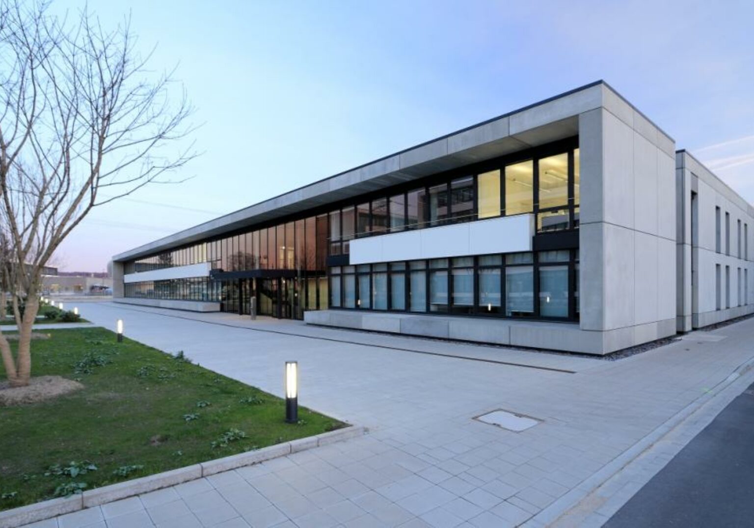 Moderne Bürogebäude-Fassade bei Sonnenuntergang mit Glasfenstern und gepflastertem Eingang, umgeben von Grünflächen.