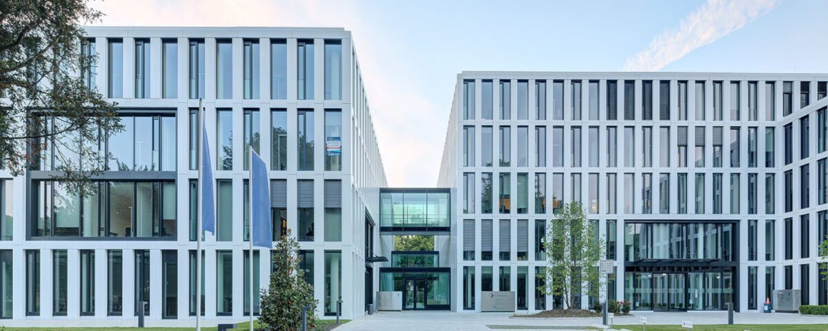 Moderne Bürogebäude mit Glasfronten, umgeben von Grünflächen und Gehweg im Vordergrund.