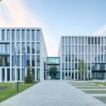 Moderne Bürogebäude mit Glasfronten, umgeben von Grünflächen und Gehweg im Vordergrund.