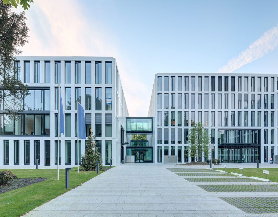 Moderne Bürogebäude mit Glasfronten, umgeben von Grünflächen und Gehweg im Vordergrund.