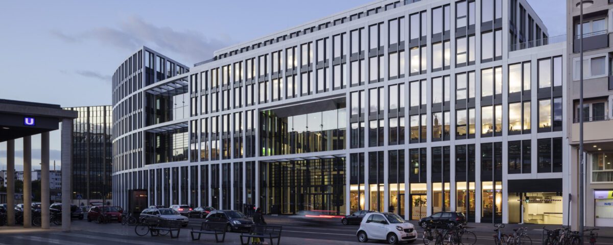 Moderne Bürogebäude neben U-Bahnstation am Abend mit parkenden Autos und Fahrrädern, Himmel wird langsam dunkel.