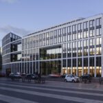 Moderne Bürogebäude neben U-Bahnstation am Abend mit parkenden Autos und Fahrrädern, Himmel wird langsam dunkel.