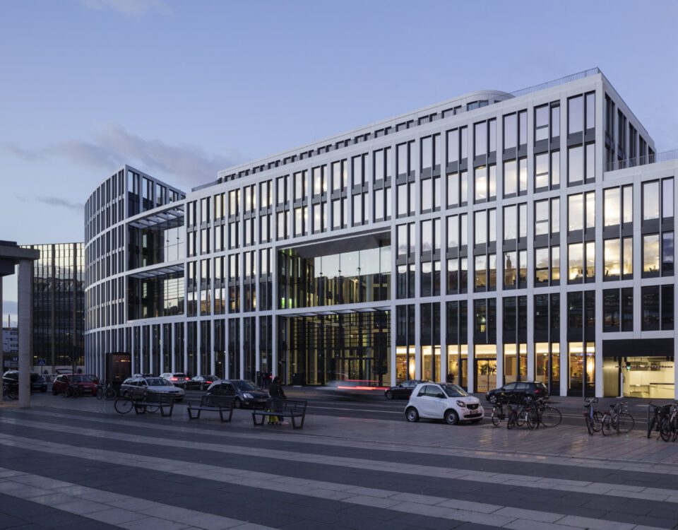 Moderne Bürogebäude neben U-Bahnstation am Abend mit parkenden Autos und Fahrrädern, Himmel wird langsam dunkel.
