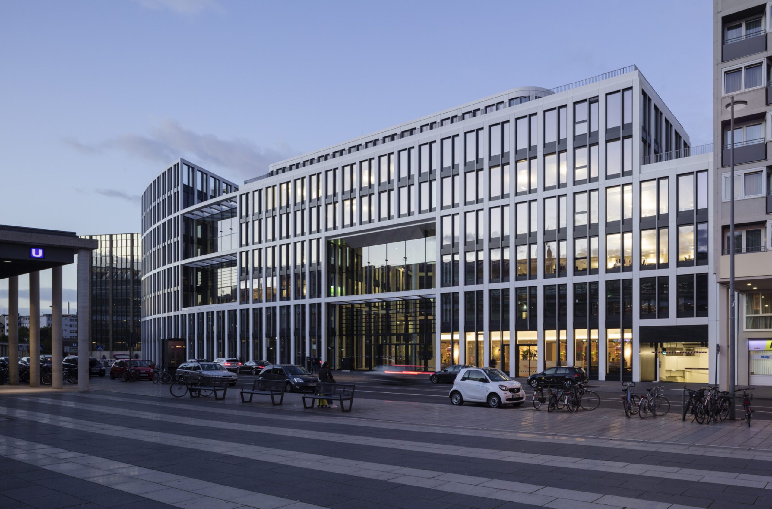 Moderne Bürogebäude neben U-Bahnstation am Abend mit parkenden Autos und Fahrrädern, Himmel wird langsam dunkel.