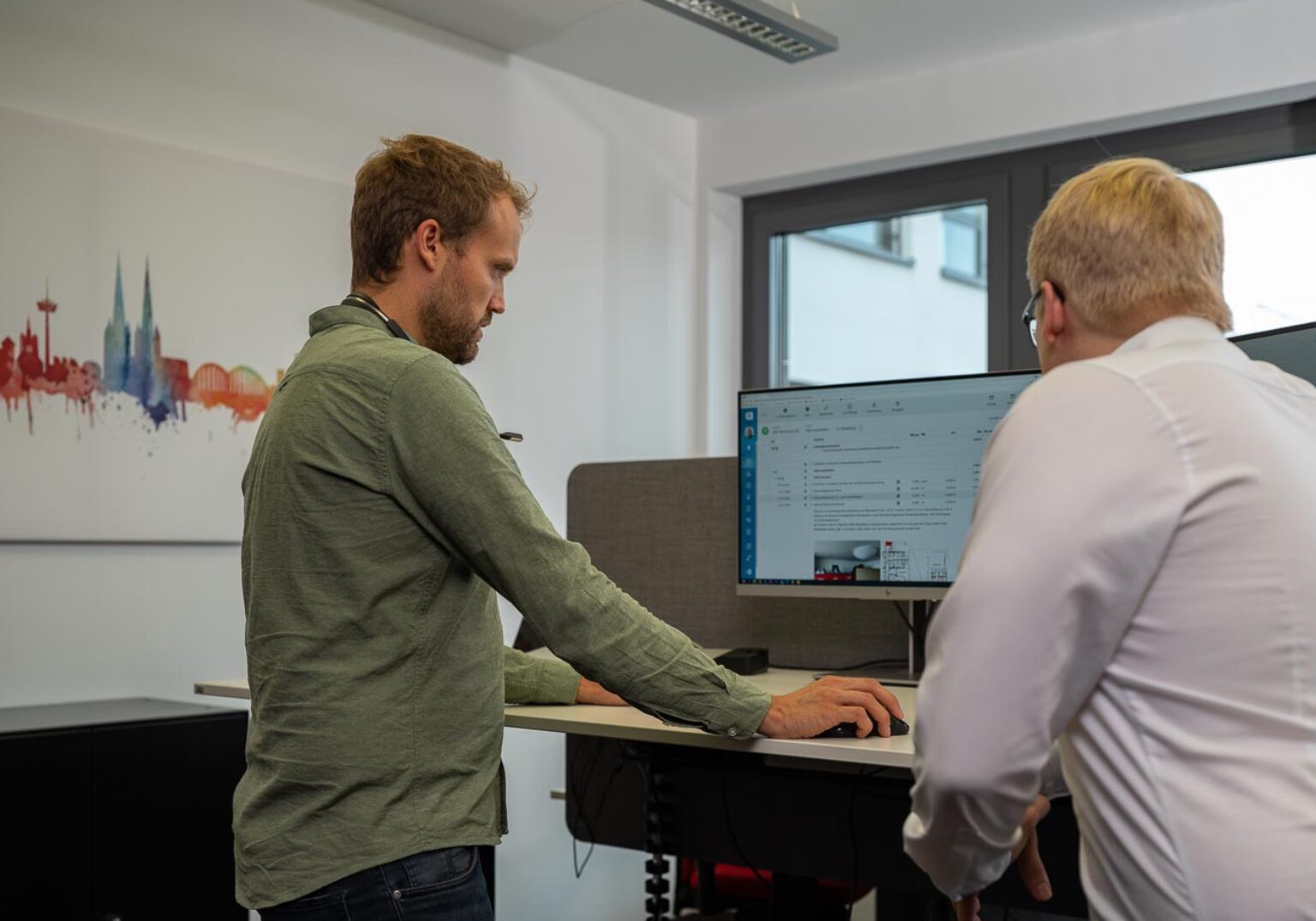 Zwei Männer arbeiten am Computer im Büro, Bild von Kölner Skyline im Hintergrund.