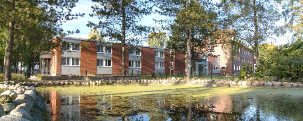 Roter Backsteinbau am Teich, umgeben von Bäumen und spiegelndem Wasser unter klarem Himmel.