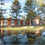 Roter Backsteinbau am Teich, umgeben von Bäumen und spiegelndem Wasser unter klarem Himmel.