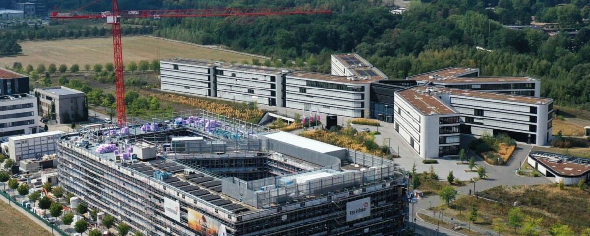 Luftaufnahme eines modernen Bürokomplexes mit im Bau befindlichen Gebäuden, umgeben von Natur und Grünflächen.