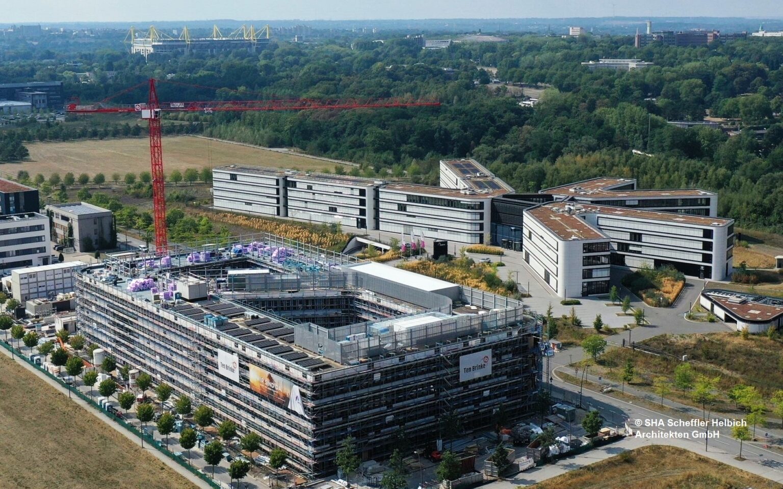 Luftaufnahme eines modernen Bürokomplexes mit im Bau befindlichen Gebäuden, umgeben von Natur und Grünflächen.