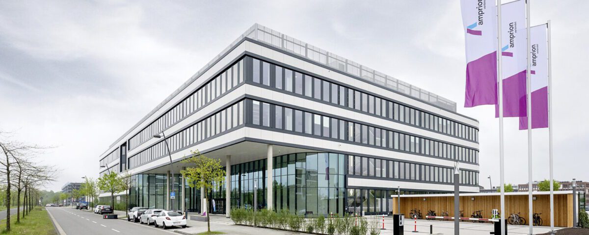Modernes Bürogebäude mit Glasfassade und Flaggen, an einer ruhigen Straße mit Autos und Bäumen gelegen.