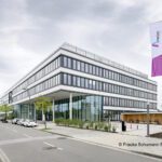 Modernes Bürogebäude mit Glasfassade und Flaggen, an einer ruhigen Straße mit Autos und Bäumen gelegen.