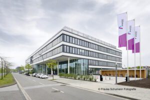 Modernes Bürogebäude mit Glasfassade und Flaggen, an einer ruhigen Straße mit Autos und Bäumen gelegen.