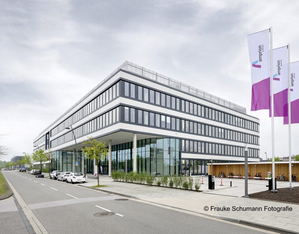 Modernes Bürogebäude mit Glasfassade und Flaggen, an einer ruhigen Straße mit Autos und Bäumen gelegen.