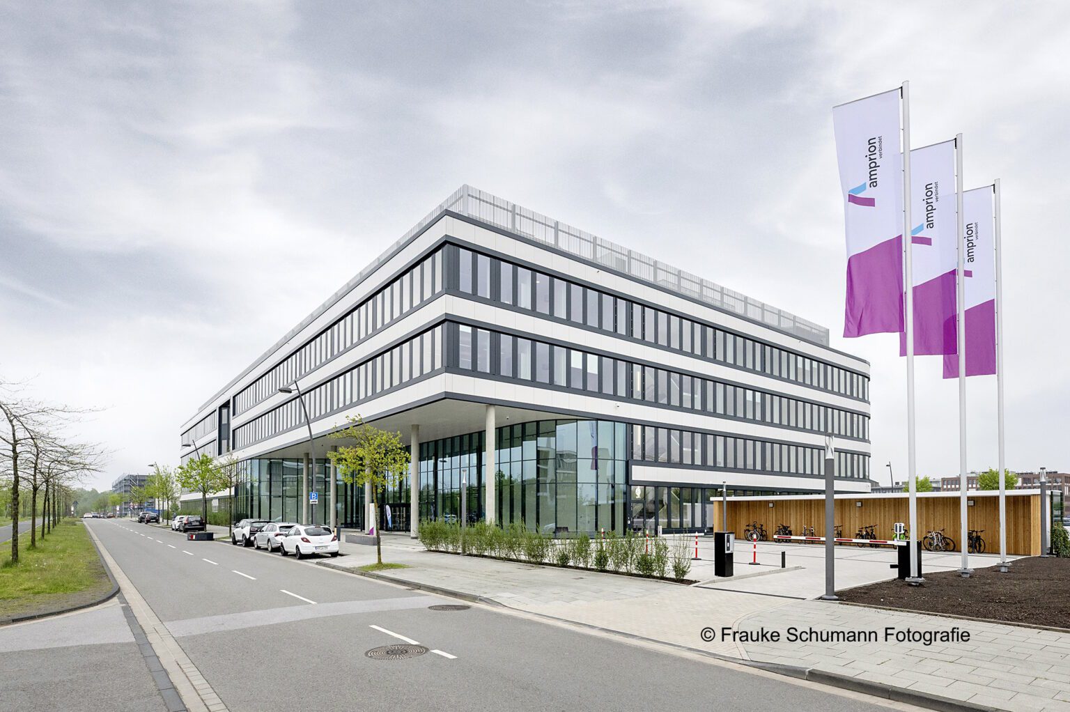 Modernes Bürogebäude mit Glasfassade und Flaggen, an einer ruhigen Straße mit Autos und Bäumen gelegen.