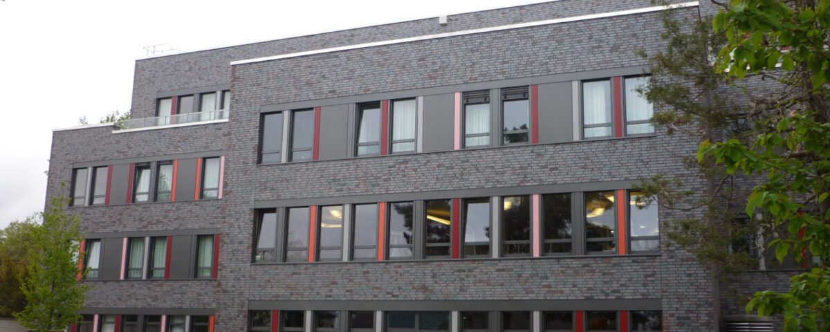 Moderne Fassade eines mehrstöckigen Bürogebäudes mit großen Fenstern, Terrasse und umgebenden Bäumen.