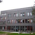 Moderne Fassade eines mehrstöckigen Bürogebäudes mit großen Fenstern, Terrasse und umgebenden Bäumen.
