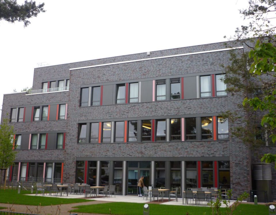 Moderne Fassade eines mehrstöckigen Bürogebäudes mit großen Fenstern, Terrasse und umgebenden Bäumen.
