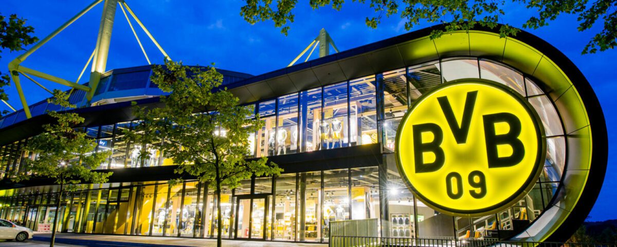 Borussia Dortmund Stadion bei Nacht, leuchtendes BVB-Logo, modernes Design, beleuchtete Glasfront und markante Architektur.