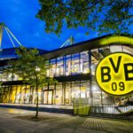 Borussia Dortmund Stadion bei Nacht, leuchtendes BVB-Logo, modernes Design, beleuchtete Glasfront und markante Architektur.