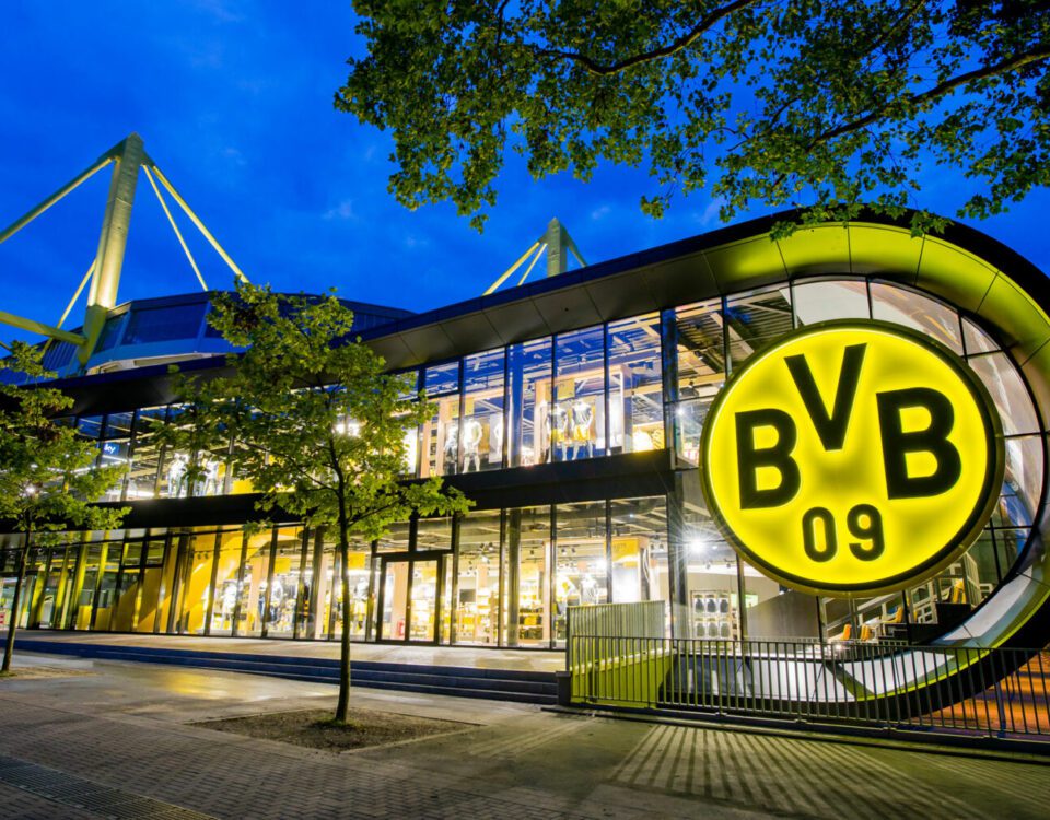 Borussia Dortmund Stadion bei Nacht, leuchtendes BVB-Logo, modernes Design, beleuchtete Glasfront und markante Architektur.